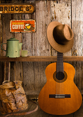  A guitar leans against a weathered bench and a well traveled  back pack sits next to it A fedora hangs on the head of the guitar. A green enamel coffee pot sits on the bench. 